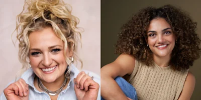 Two women smiling; one with blonde curly hair in a bun wearing a light shirt, the other with dark curly hair wearing a beige sleeveless top.