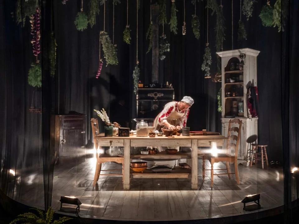 A person in a chef’s hat and apron prepares food at a wooden table on a stage set resembling a rustic kitchen, with herbs hanging from above and shelves in the background.