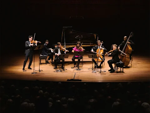 Seven musicians perform onstage with a variety of string instruments and a piano, facing an audience in a concert hall.