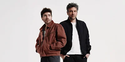 Two men stand side by side against a plain white background, one wearing a brown jacket and the other in a black jacket over a white shirt.