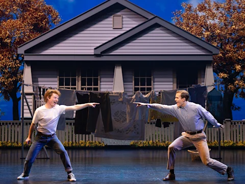 Two people are dancing and pointing at each other on stage in front of a house backdrop with laundry hanging on a clothesline.