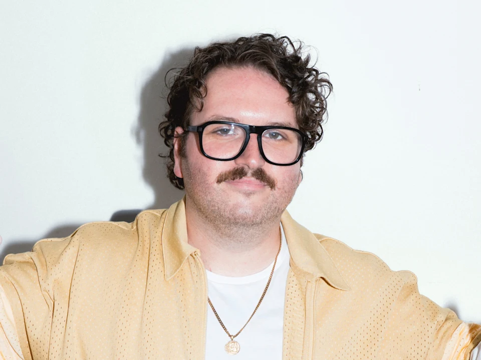 A man with curly hair, glasses, and a mustache wears a light yellow shirt over a white t-shirt, standing against a plain white background.
