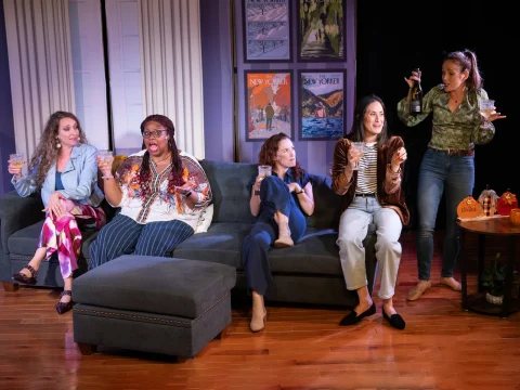 Five women are gathered in a living room; three are sitting on a sectional sofa, one is sitting on the floor, and one is standing holding two drinks. They all appear to be engaged in conversation.