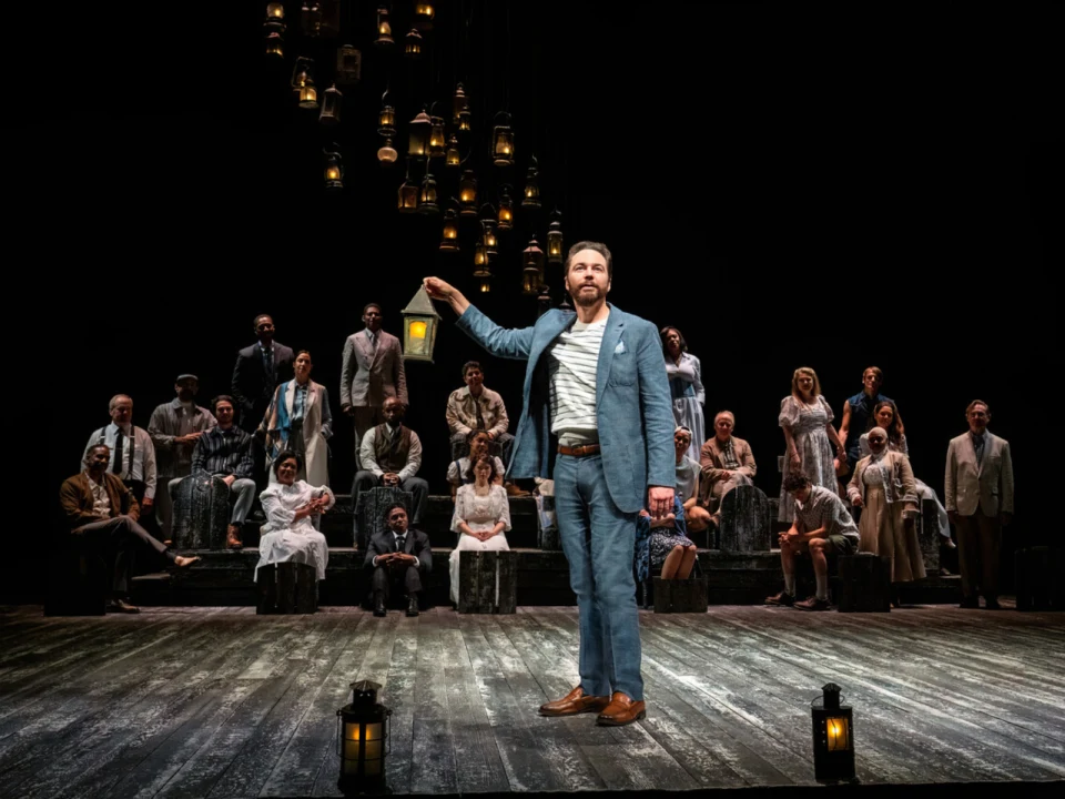 A man in a blue suit holds a lantern on a dimly lit stage. Behind him are rows of people sitting and standing on steps, with hanging lanterns above.