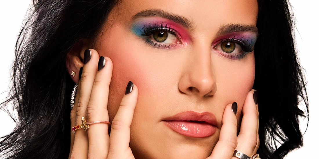 Close-up of a woman with colorful eyeshadow, glossy lips, manicured nails, multiple rings, and earrings, holding her face with both hands against a white background.