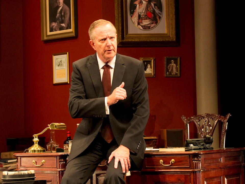 A man in a suit sits on a desk in an office setting with framed portraits on a red wall behind him, gesturing with his right hand.