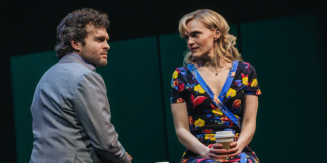 A man in a gray suit and a woman in a floral dress sit facing each other on stage. The woman holds a coffee cup and both have serious expressions.