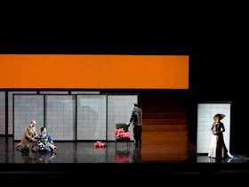Four actors perform on a minimalist stage with white panels and an orange backdrop; two are kneeling by a woman, one stands by a chair with pink flowers, and one stands alone at right.