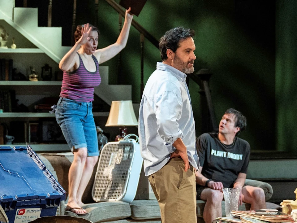 Three adults are in a cluttered living room; a woman stands on a couch raising her arms, while two men watch her, one sitting and one standing with his back partially turned.