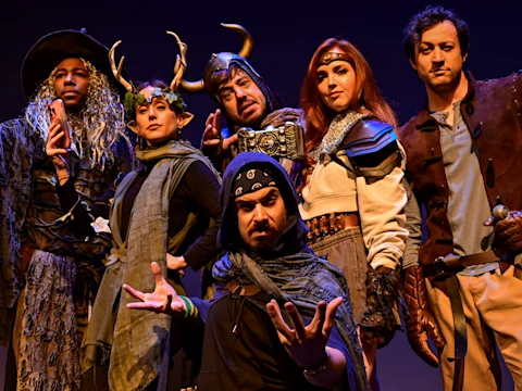 Six adults in elaborate fantasy costumes pose together against a dark background, dressed as characters including a wizard, a druid, a warrior, and a rogue.