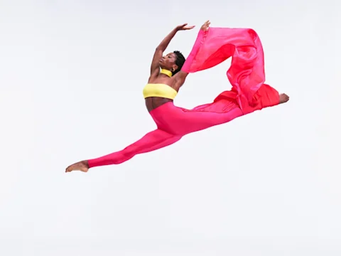 Dancer in a yellow top and pink pants mid-air with arms outstretched and a flowing pink fabric behind against a white background.