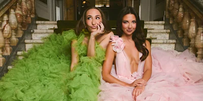 Two women sit on marble stairs wearing elaborate ruffled gowns; one in green with a playful expression, the other in pink with a calm demeanor.