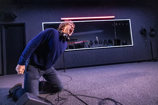 A man with headphones around his neck kneeling and shouting in a recording studio.