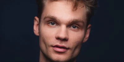 A young adult with light skin and short, tousled brown hair looks directly at the camera against a dark background.