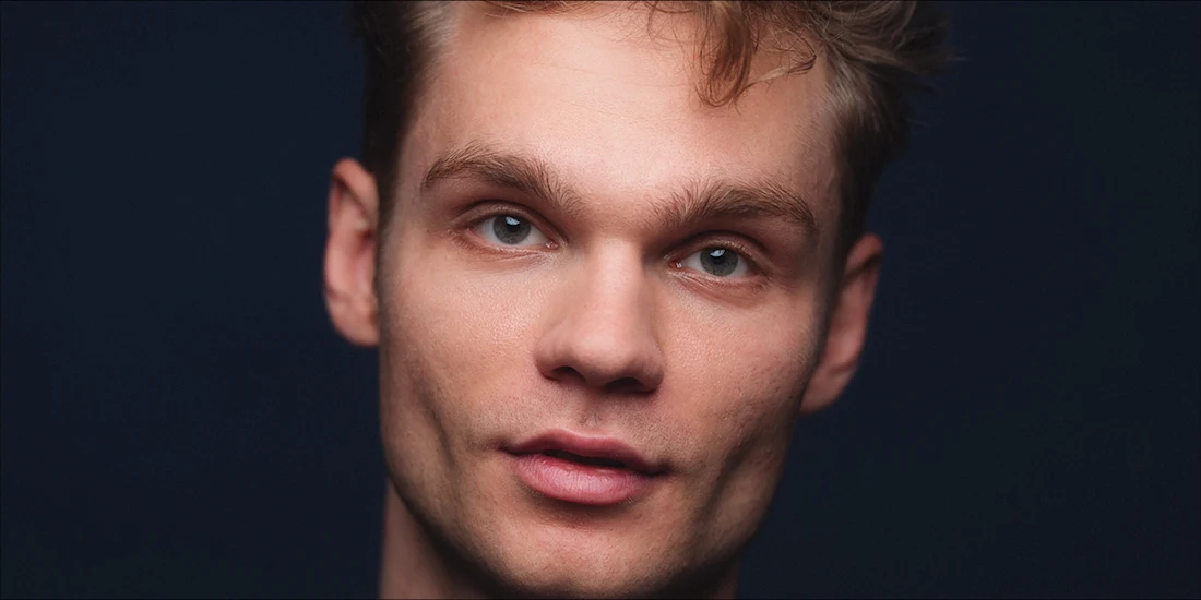 A young adult with light skin and short, tousled brown hair looks directly at the camera against a dark background.
