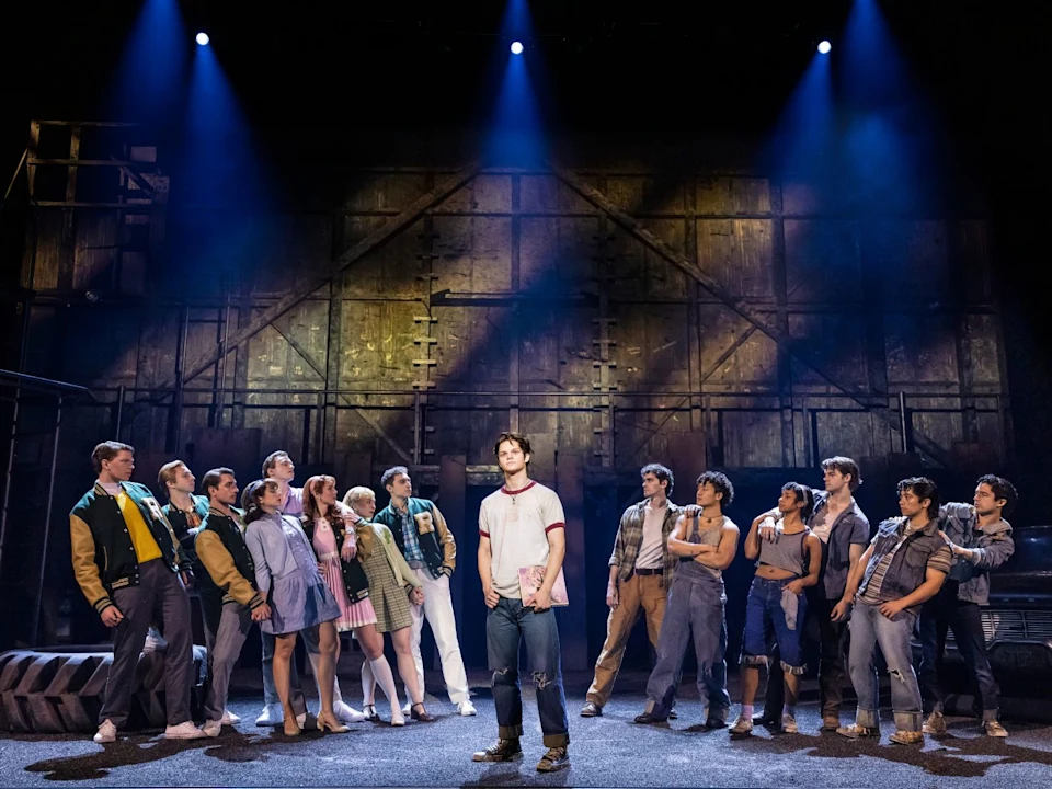 A group of actors stand in a semicircle on stage facing a single performer in the center, with dramatic lighting and an industrial set in the background.