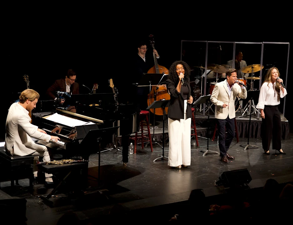 Six musicians perform on stage; one plays piano, one plays double bass, one plays guitar, one plays drums, and three vocalists sing into microphones.