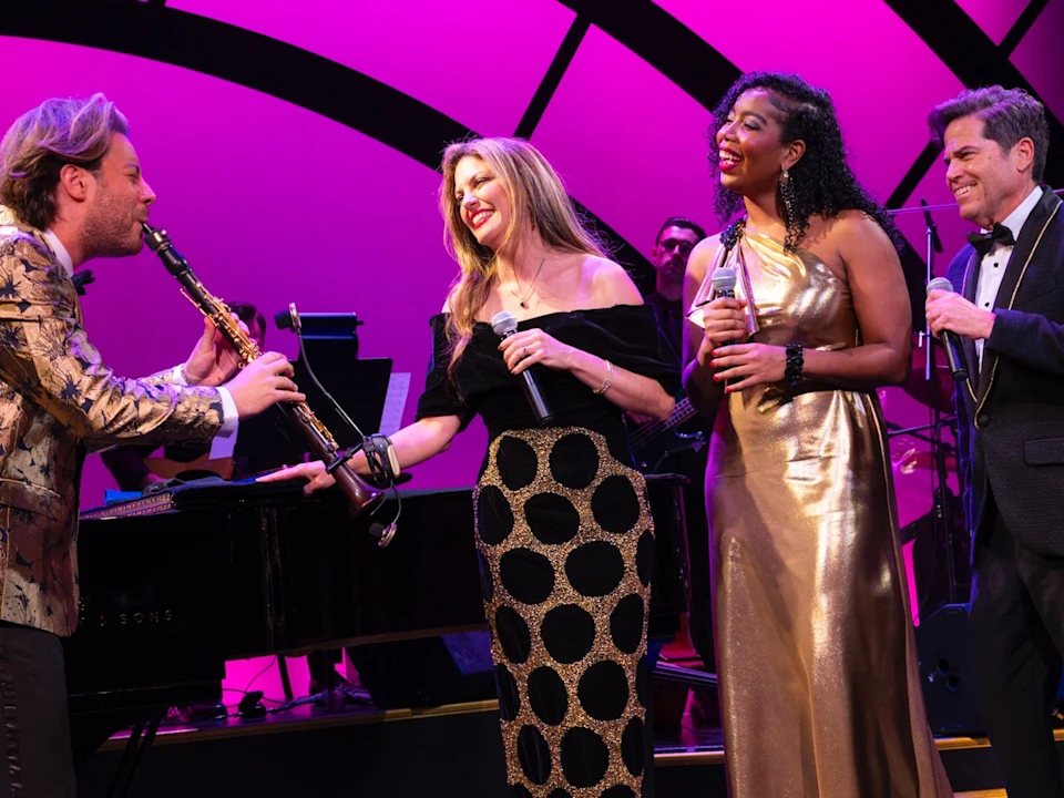 Four musicians perform on stage under pink lighting. Three are singing into microphones, and one plays the clarinet. Everyone is dressed in formal attire.