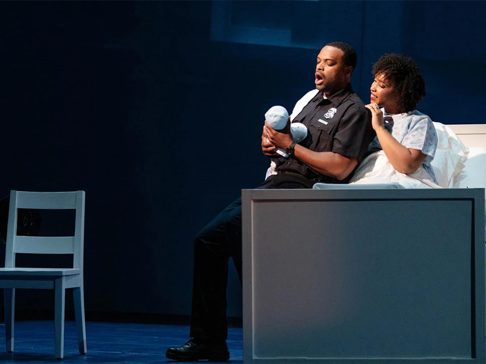 Production photo of Blue in Chicago, showing a man in a police uniform and a woman in a hospital gown sit on a bed. The man holds a small bundle while the woman looks on. A white chair is positioned nearby.