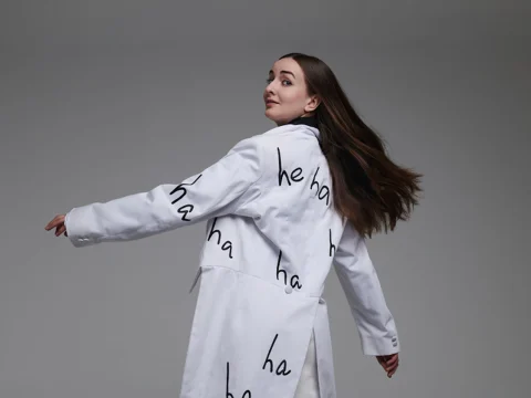A person with long hair, wearing a white coat with "ha ha ha" printed on it, turns to face the camera against a plain background.