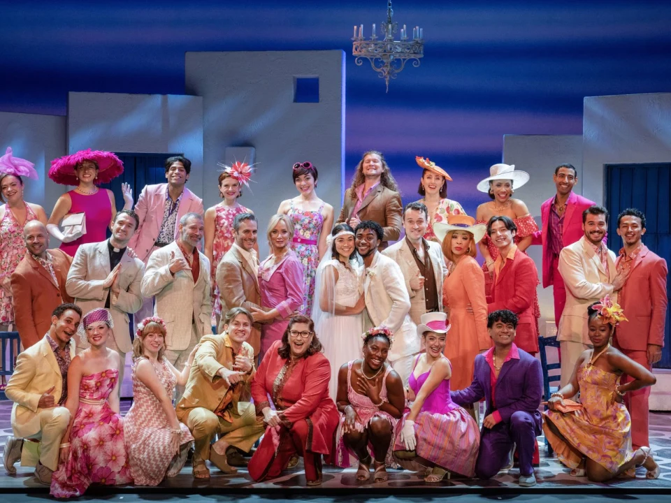 A group of people of mixed ages and genders, dressed in colorful outfits, posing together on a brightly lit stage with a Mediterranean backdrop.