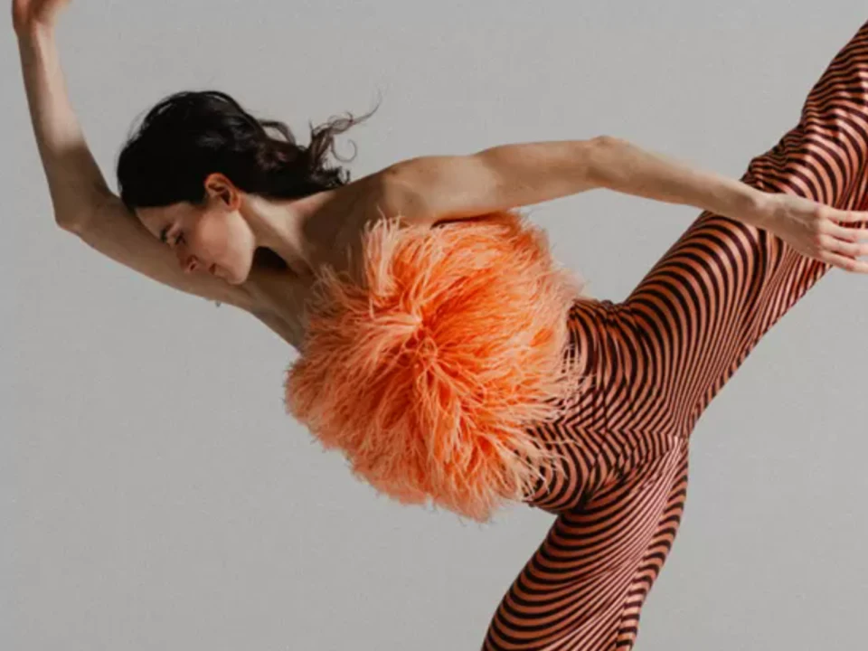 A dancer in a dynamic pose wearing an orange feathered top and striped pants on a neutral background.