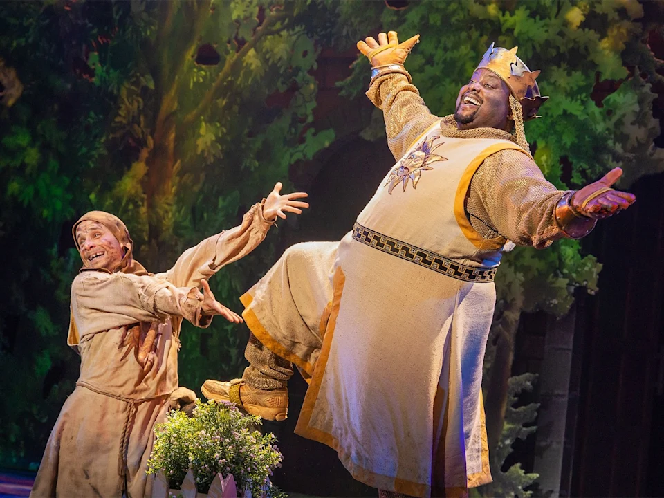 Two actors in medieval costumes perform energetically on stage; one wears a crown and regal attire, while the other is dressed in tattered clothes. Trees and foliage are visible in the background.