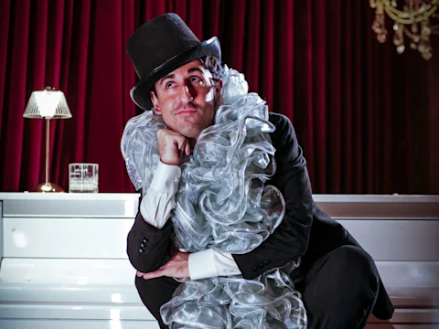 A person in a suit and top hat sits with a hand on their chin, wearing a large ruffled collar, in front of a white piano and red curtains.