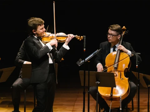 Two musicians in formal attire perform on stage. One plays the violin while standing, and the other plays the cello while seated.