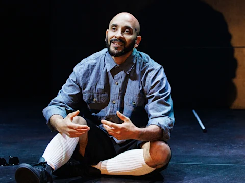 A person with a shaved head sits cross-legged on stage, gesturing with their hands, wearing a buttoned shirt, shorts, and knee-high white socks.