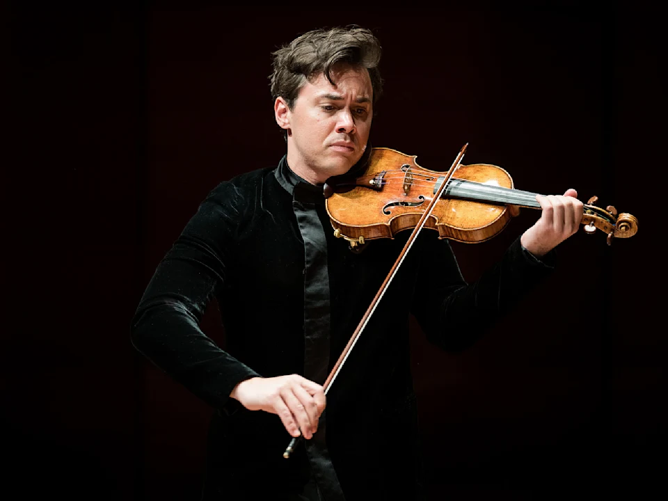 A man in a black velvet jacket plays a violin with concentration against a dark background.