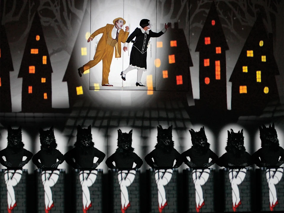 Two actors in costume perform on a stage with a backdrop of stylized houses, while a row of people in wolf masks and white pants pose below.