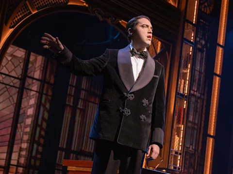Production photo of The Great Gatsby with Ryan in a black tuxedo stands on stage, gesturing with his right hand, with a decorative arch and set pieces in the background.