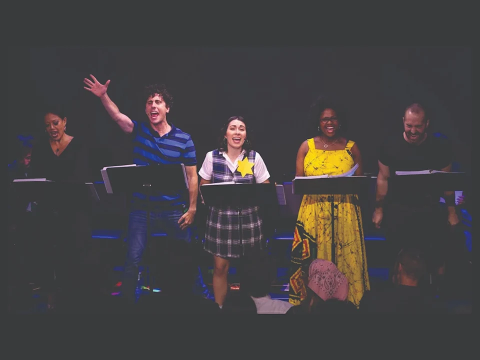 Five actors stand on stage behind music stands, performing energetically with scripts in hand, in front of an audience.