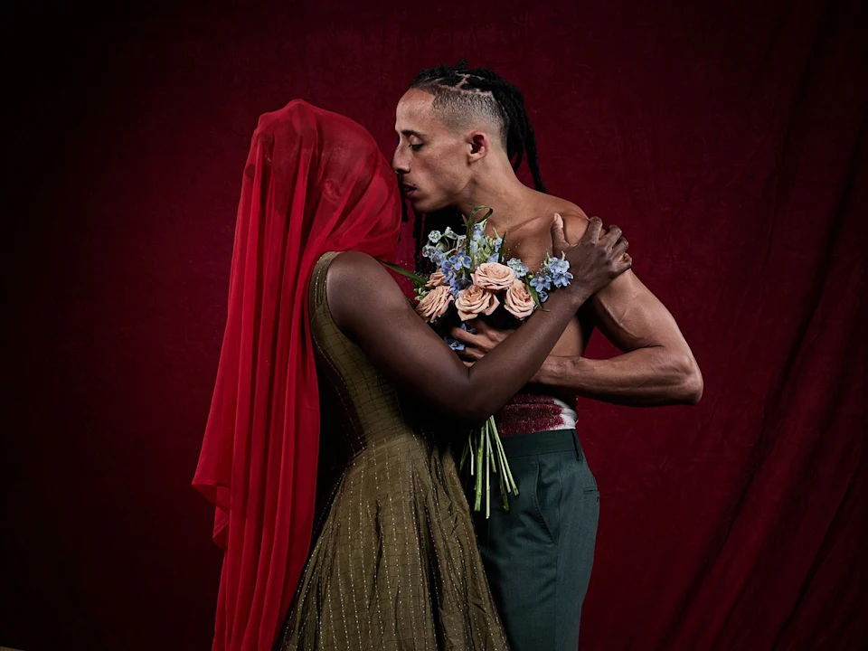 A person in a red veil embraces a shirtless person holding a bouquet of flowers against a dark red backdrop.