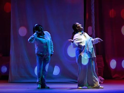 Two people in loose, light-colored clothing stand on stage under colored lights, appearing to sing or perform theatrically against a backdrop with circular light projections.