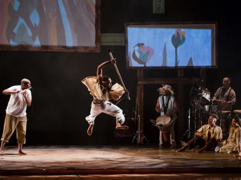 Production photo of The Migration: Reflections on Jacob Lawrence in Washington with one performer leaping mid-air with a staff, while others are playing instruments.