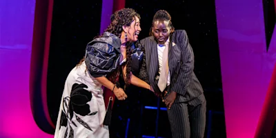 Two performers on stage: one in a black-and-white dress, the other in a pinstripe suit and tie, both smiling under pink and red stage lighting.