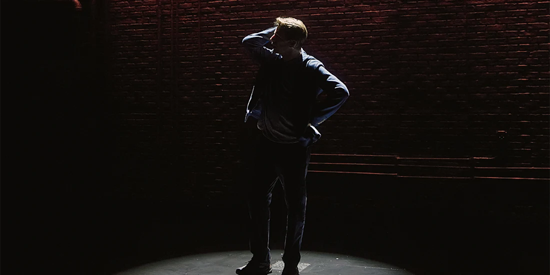A person stands alone on a dimly lit stage, silhouetted by a spotlight, with one hand on their head in front of a brick wall background.
