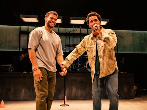 Two men stand on stage, one holding a microphone and smiling while speaking, the other standing beside him and smiling. Musical instruments and stage lighting are visible in the background.