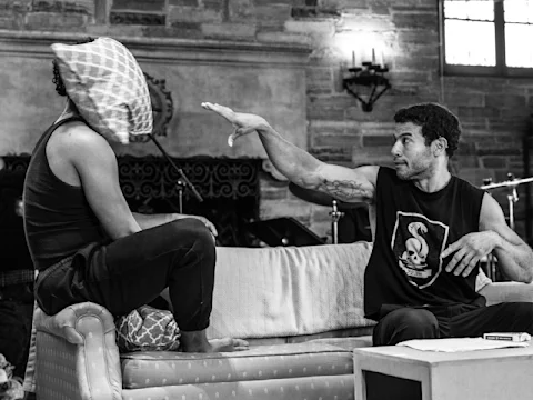 Two people sit on a couch; one has a pillow over their face while the other raises a hand as if mid-gesture. The scene appears to be an intense or dramatic conversation.