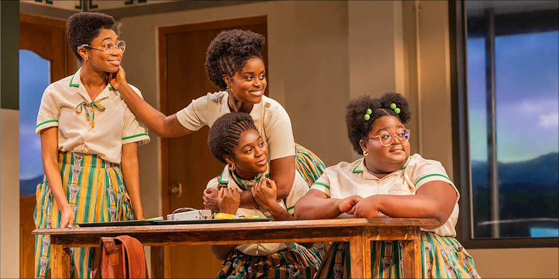 Four girls in matching school uniforms sit and stand around a wooden table, smiling and interacting in a brightly lit classroom setting with large windows in the background.