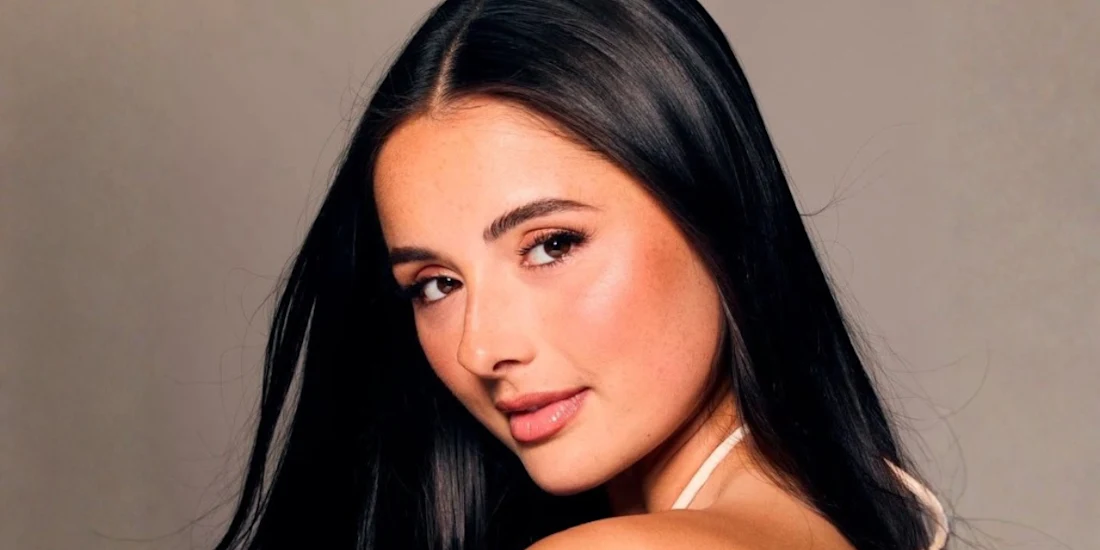 A woman with long straight dark hair poses against a neutral background, looking over her shoulder with a slight smile.