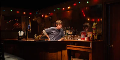 A man stands behind a wooden bar with snacks, cups, and bottles; colored string lights and a deer head are on the wall behind him.