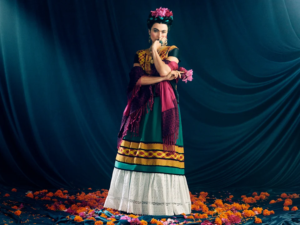 Production photo of El Último Sueño de Frida y Diego in New York, showing person in traditional Mexican dress with floral headpiece stands on a dark stage, surrounded by orange marigold petals, with a thoughtful pose and dark draped fabric backdrop.