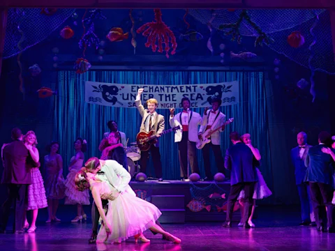 Performers on stage reenact a 1950s high school dance with a live band under a banner reading "Enchantment Under the Sea," as couples dance and one couple shares a dramatic kiss.