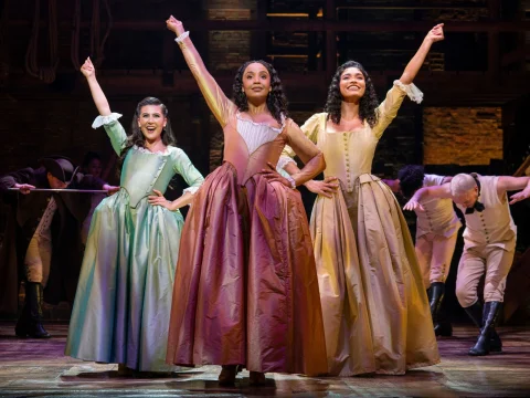 Three women in period costumes strike a pose on stage with raised arms and smiling faces. The background features a wooden set with performers in colonial-style clothing.