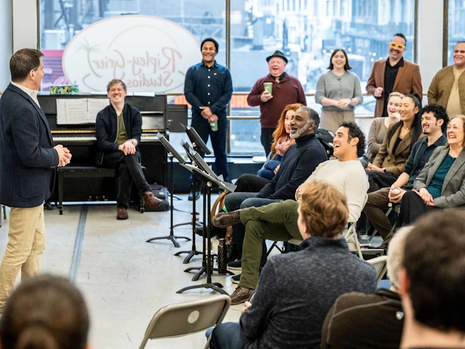 A group of people sit and stand in a studio, listening to a speaker; some are smiling or laughing, with music stands and a piano visible in the background.