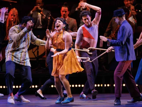 People dancing energetically on stage, one woman in an orange dress is in the center, surrounded by others in colorful outfits. A band is visible in the background.