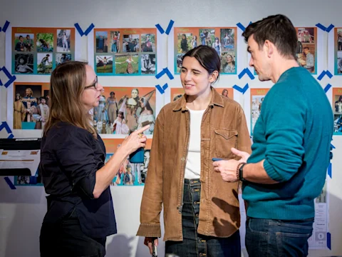 Three people are having a discussion in front of a wall covered with photos and notes arranged in a grid pattern, some marked with blue tape.